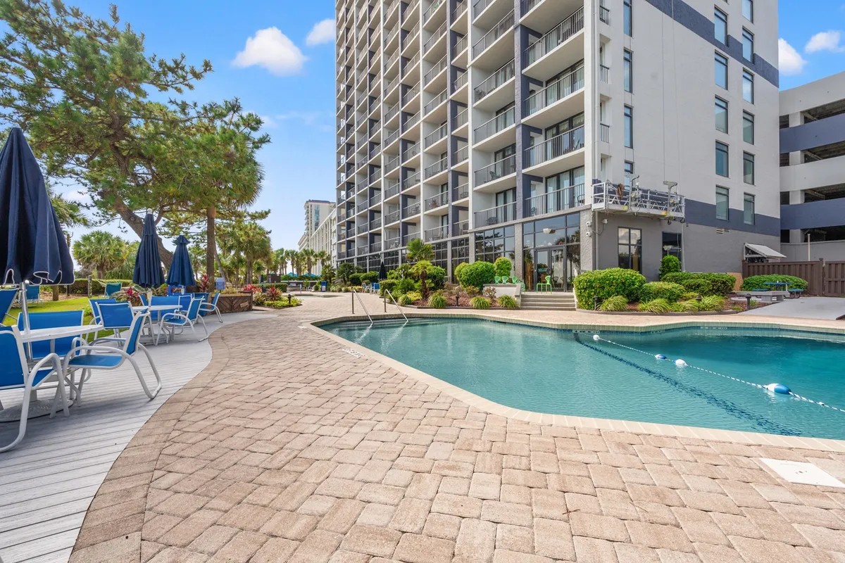 A lively resort pool scene with sparkling turquoise water, plenty of lounge chairs, and lush greenery surrounding the building's facade. The scene feels energetic and social, perfect for guests seeking fun on property—whether swimming, sunbathing, or gathering poolside.
