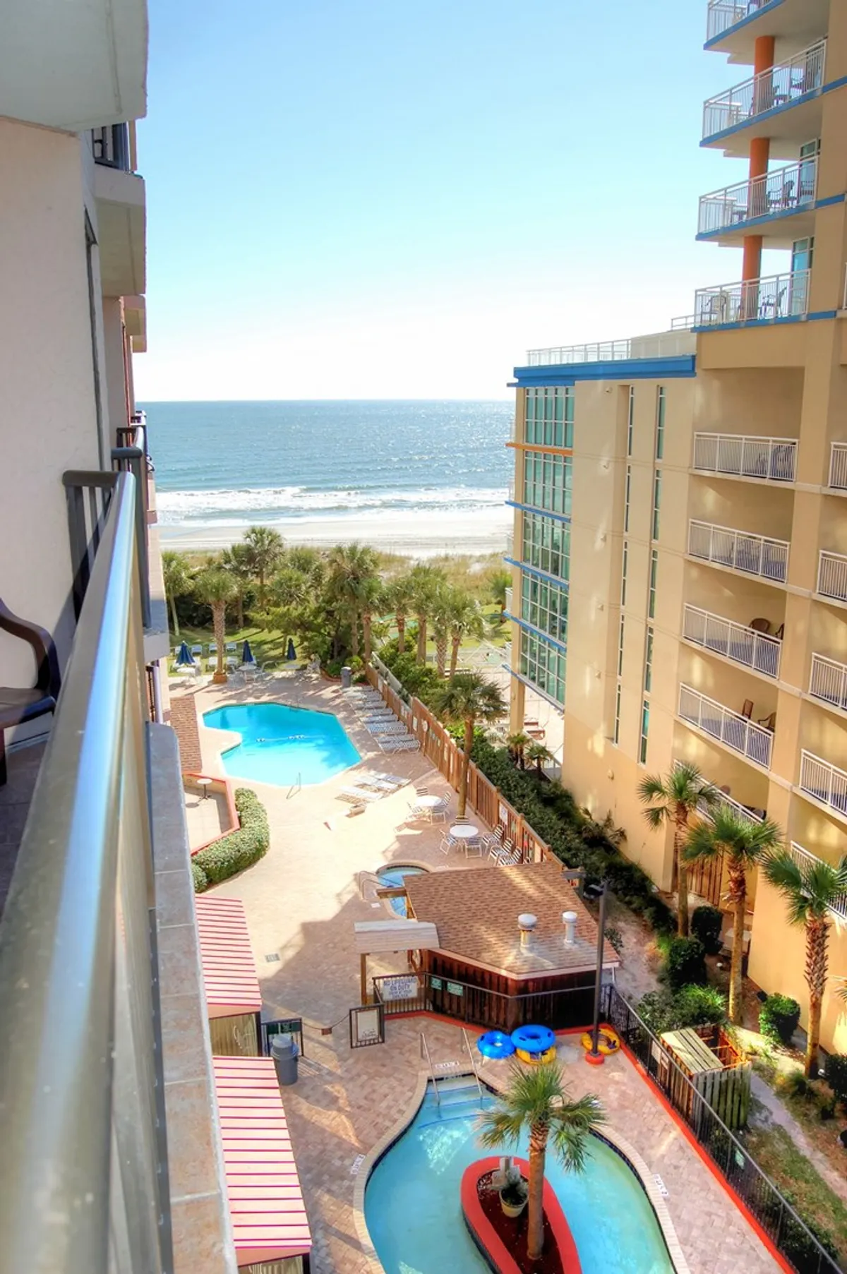 A stunning elevated view from a resort balcony overlooking multiple swimming pools, palm trees, and the vast ocean beyond. The scene exudes a vacation vibe, inviting families to relax, swim, and socialize after a day of exploring local attractions.