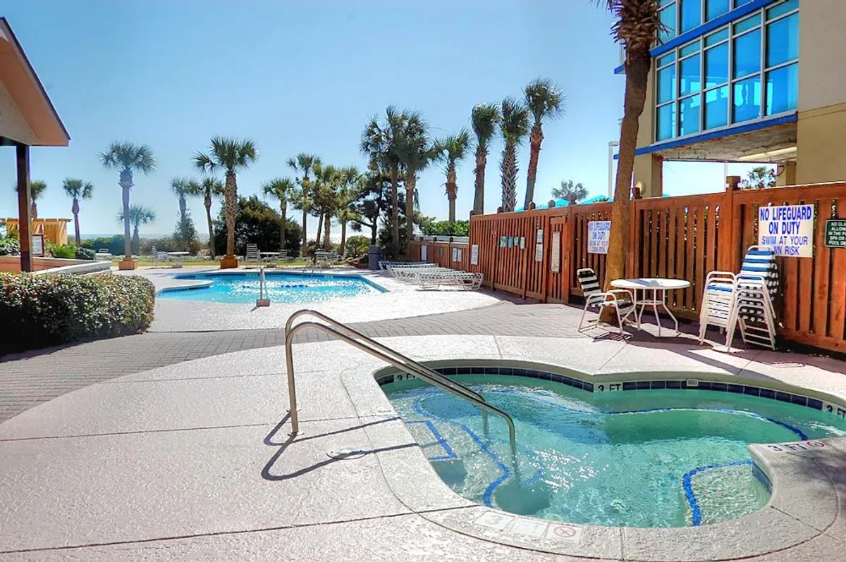The scene features a spacious outdoor swimming pool with an adjacent jacuzzi, plenty of lounge chairs, and inviting palm trees creating a true vacation vibe. The area is designed for both swimming and relaxing in the hot tub, with ample seating for socializing.