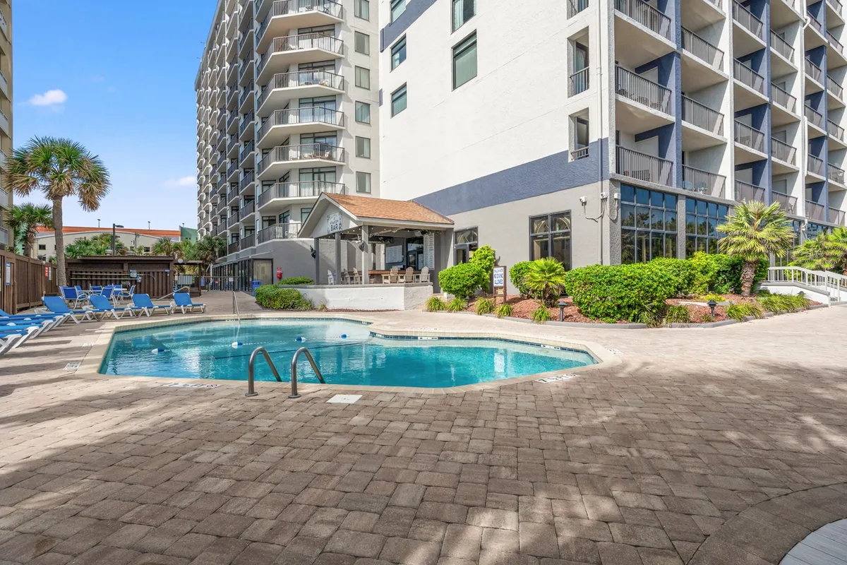 A sunlit outdoor swimming pool surrounded by lounge chairs, landscaped greenery, and a covered gazebo seating area. The poolside environment suggests a leisurely, social, and relaxing atmosphere—ideal for extended stays in mild climates.