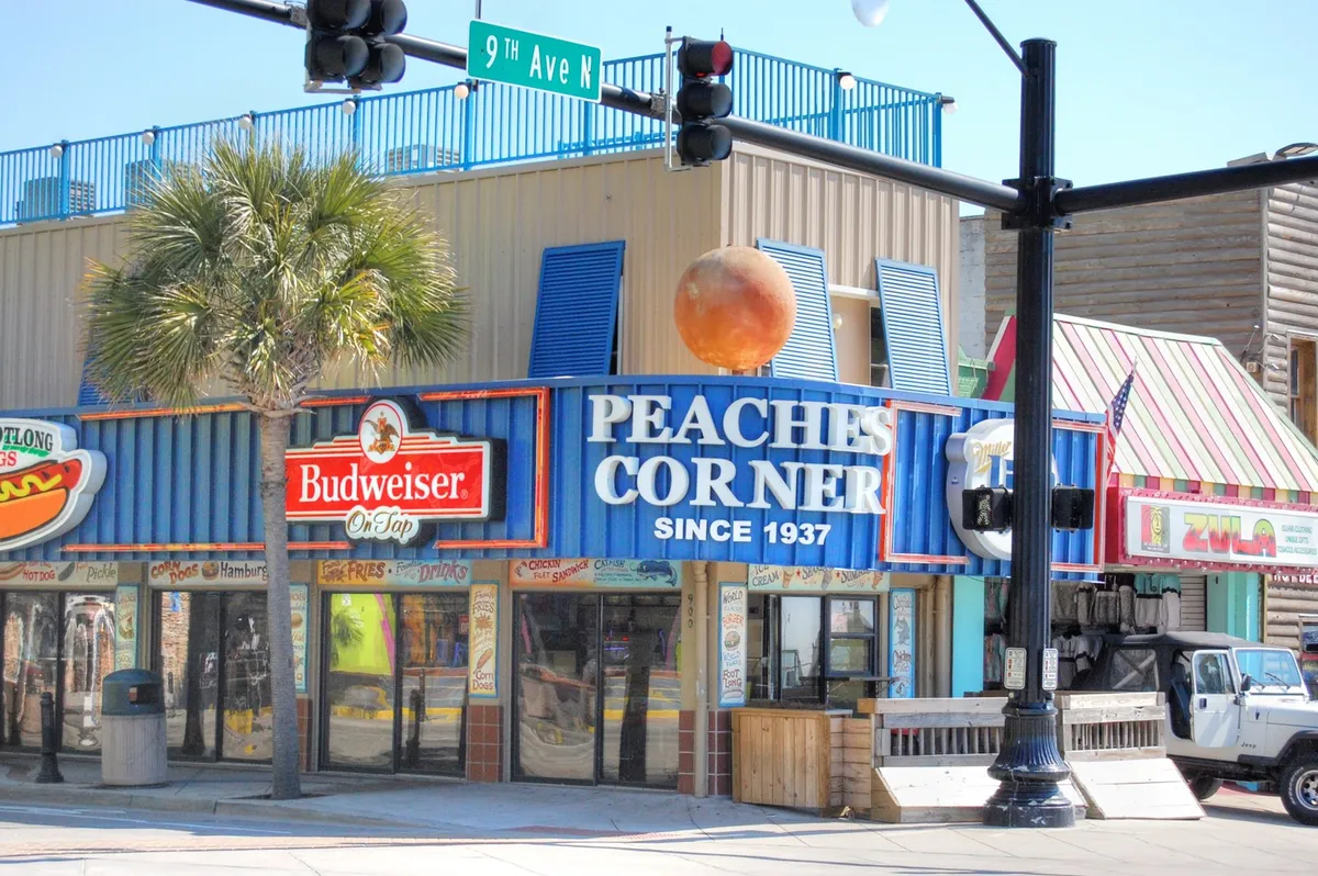 A colorful street scene with the Peaches Corner and Budweiser signs, a palm tree, and local traffic, suggesting a festive atmosphere filled with dining, music, and socializing opportunities—hallmarks of Myrtle Beach’s nightlife and local flavor.