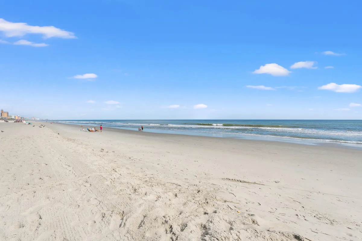 A tranquil stretch of sandy beach meets gentle ocean waves, with a few people strolling along the shoreline. The sky is clear, and the atmosphere is peaceful, evoking relaxation and romance.