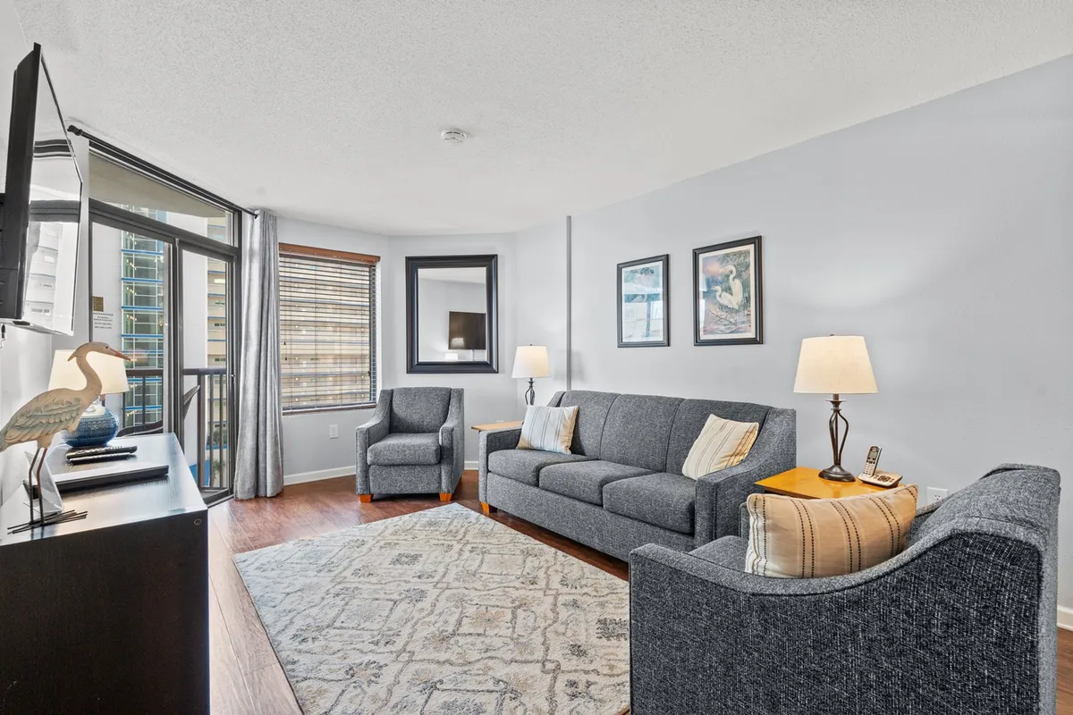 The image shows a stylish, inviting living room with a plush sofa, cozy armchairs, a large TV, soft lighting from a table lamp, and vibrant artwork. The space is designed for relaxation and quality time, offering comfort and modern amenities.