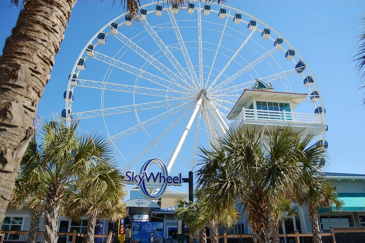 This image features the iconic Myrtle Beach SkyWheel, framed by palm trees and a bustling ticket booth, setting a joyful, amusement-filled atmosphere. The scene captures the excitement of a classic boardwalk attraction that appeals to families seeking memorable experiences.