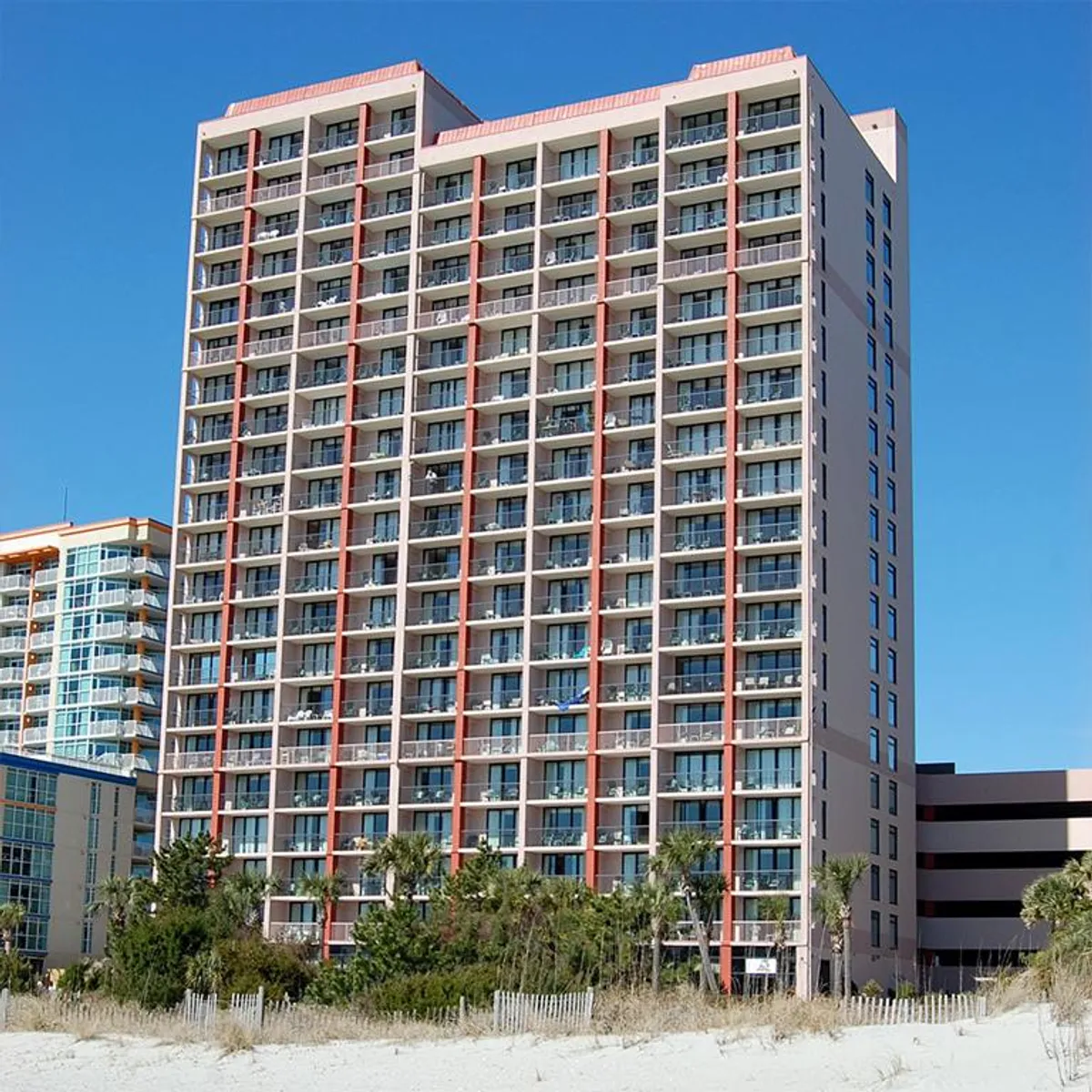 The tall facade of Beach Colony Resort, accented by palm trees and lush landscaping, stands near the beach. Numerous balconies suggest private spaces for taking in the view, while the relaxed coastal setting encourages both rest and exploration.