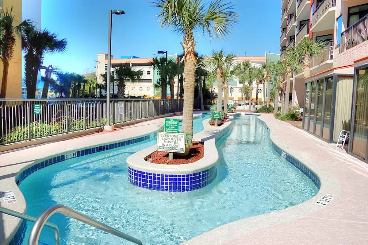 This image showcases the resort's lazy river pool winding through a tropical setting with palm trees and visible signage. Lounge chairs line the area, suggesting a fun and leisurely environment for guests of all ages.