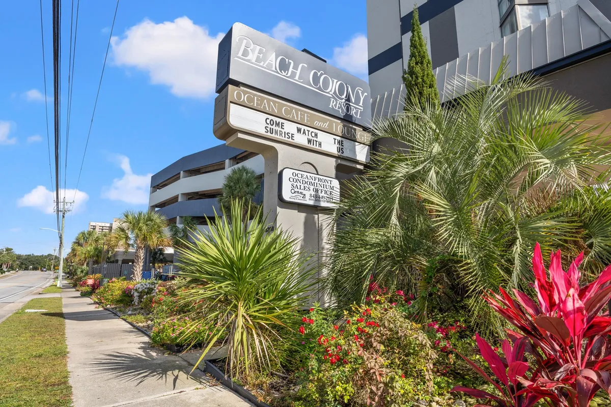 The Beach Colony Resort sign stands amid lush tropical plants and colorful flowers, with signage for the Ocean Cafe and Lounge visible. The approach is inviting and hints at on-site dining and relaxation areas.